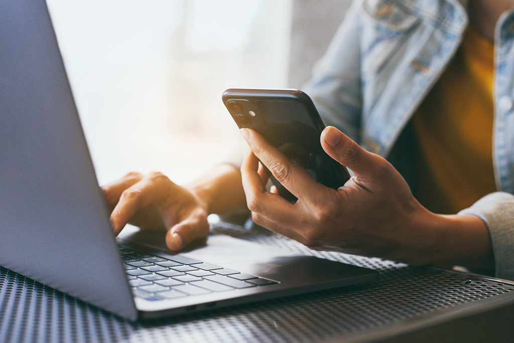 A person using a laptop and holding a phone